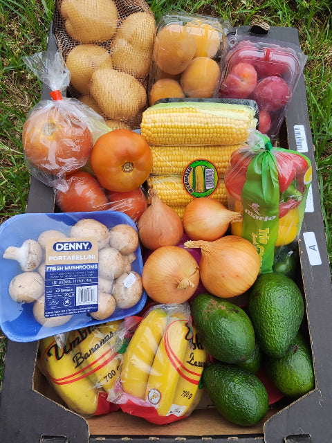 Assorted fresh produce including corn, tomatoes, avocados, and mushrooms in a cardboard box outdoors.